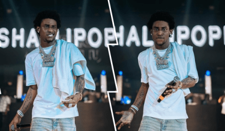 Nigerian singer Shallipopi performing on stage. He's wearing a white t-shirt, light blue jeans, and a large, ornate silver necklace. He's holding a microphone and appears to be in the midst of a performance, with a blurred background suggesting a concert setting and his name displayed prominently behind him.