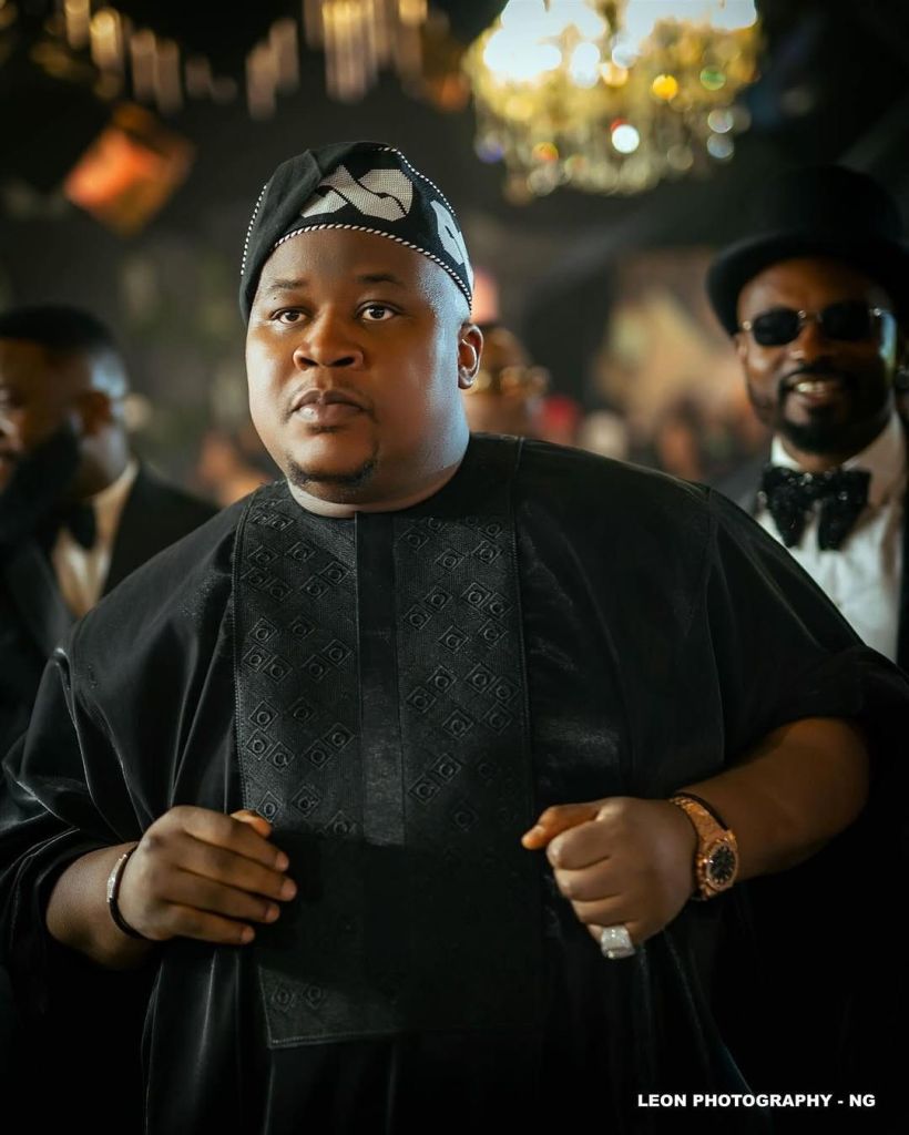 Cubana Chief Priest stands in a luxurious setting with chandeliers, dressed in a black traditional outfit with a patterned cap, accessorized with a watch and bracelet, as others in formal attire are visible in the background. Credit: Leon Photography NG.