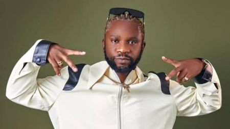 Rapper Speed Darlington poses against a green background, wearing a white jacket with black leather accents, sunglasses on his head, and gold jewelry, pointing both hands outward in a confident gesture.