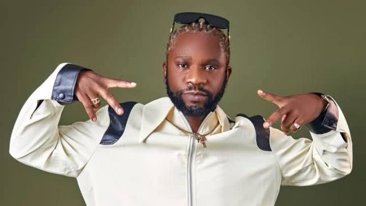 Rapper Speed Darlington poses against a green background, wearing a white jacket with black leather accents, sunglasses on his head, and gold jewelry, pointing both hands outward in a confident gesture.