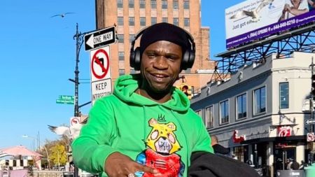 Speed Darlington stands outdoors in a vibrant green hoodie featuring a colorful cartoon bear design, wearing black headphones and a beanie, with a city street featuring a "One Way" sign and buildings in the background.