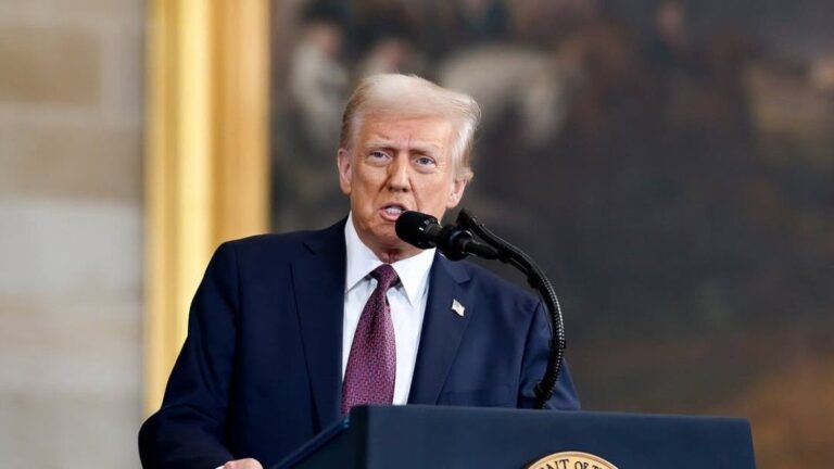 President Donald Trump speaking at a podium with the seal of the President of the United States, wearing a dark suit, white shirt, and red tie, with a blurred background featuring gold columns and a historical painting.