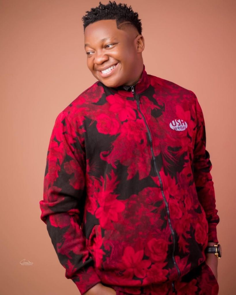 Skitmaker Mama Uka is seen wearing a white shirt with a black design, holding a photo of himself dressed in a pink top and a colorful patterned skirt. The main setting features a room with wooden elements and a window, while the inset photo presents an outdoor backdrop with buildings and a red car, highlighting his comedic versatility.