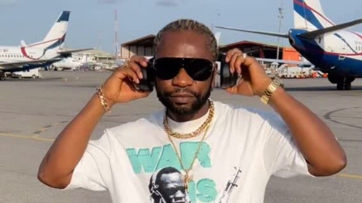 Rapper Speed Darlington stands on an airport tarmac, wearing sunglasses, a white T-shirt with "Warris" text, and multiple gold chains, while holding his headphones to his ears with both hands, with airplanes visible in the background.