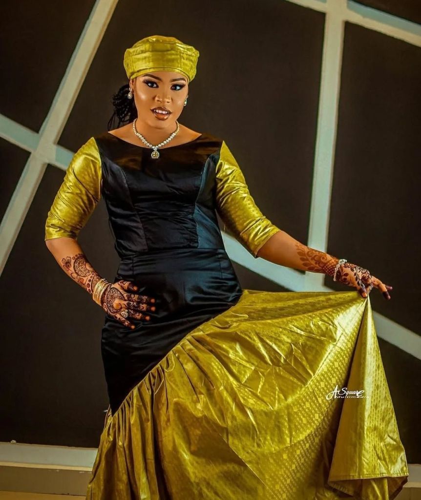 Tiktok star Murja Kunya stands elegantly in a striking black and gold dress, featuring a fitted bodice and flowing skirt. She accessorizes with a matching gold headwrap, intricate henna designs on her hands, and a pearl necklace, posing confidently against a geometric backdrop.