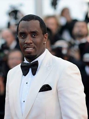 Diddy stands in a white tuxedo with a black bow tie and pocket square, surrounded by photographers at a red carpet event.