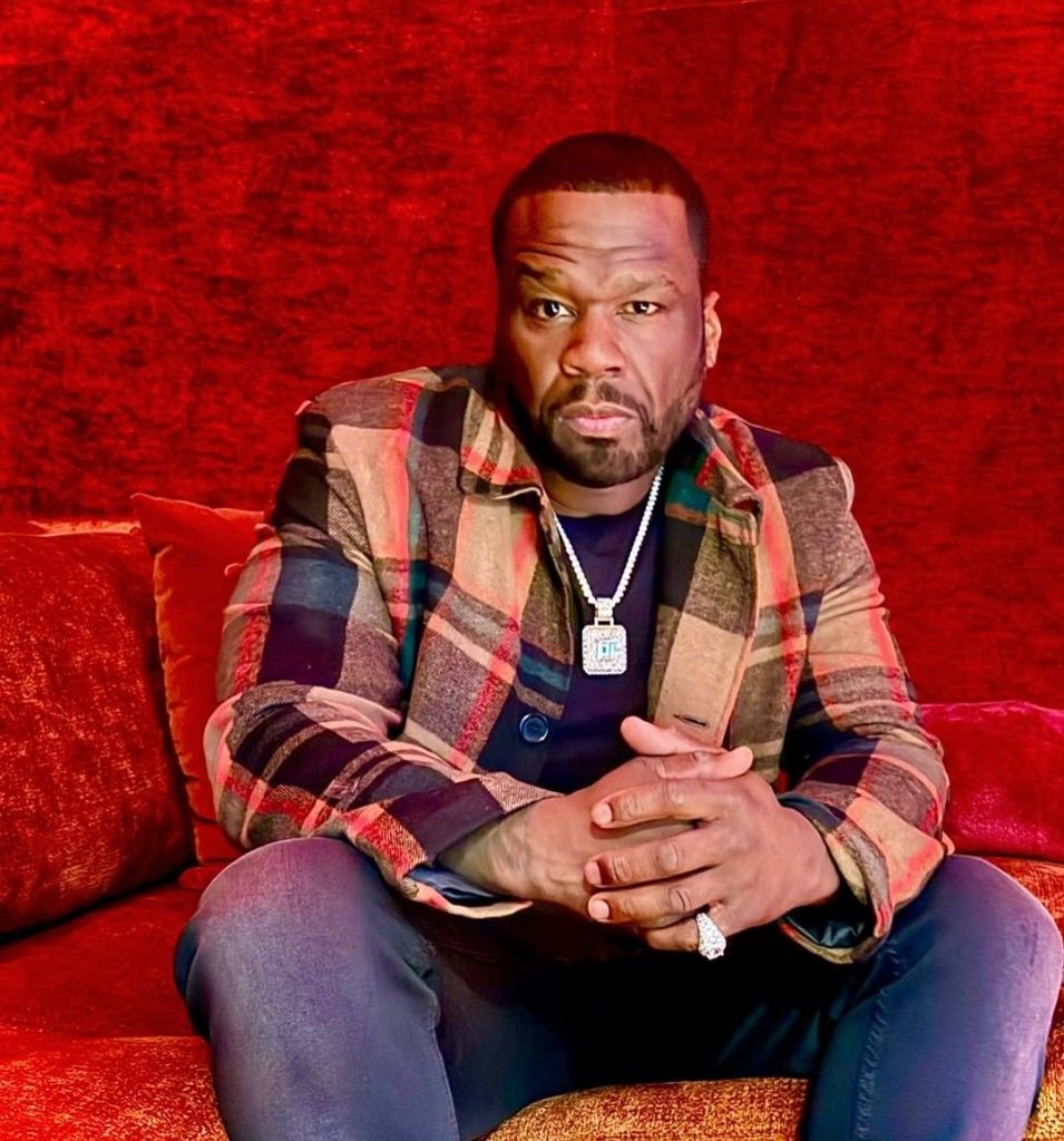 Rapper 50 Cent sits on a red couch, wearing a plaid jacket, dark shirt, and jeans, accessorized with a necklace and ring, against a matching red backdrop.
