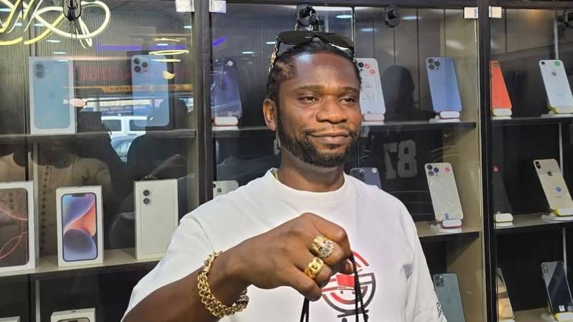Rapper Speed Darlington stands in front of a display case filled with various smartphones, wearing a white shirt with a logo, gold jewelry, and sunglasses perched on his head, pointing at the camera with a confident expression.