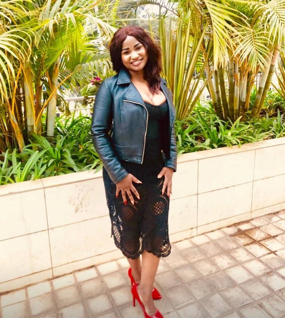 Hellen Ati stands outdoors surrounded by lush greenery, wearing a black lace dress with a leather jacket and striking red high heels.