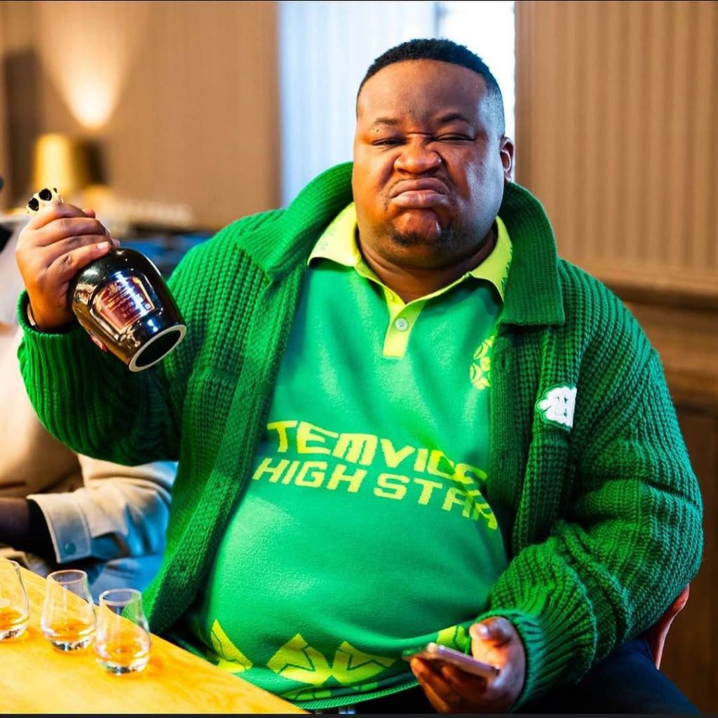 Cubana Chief Priest, dressed in a green outfit with a "High Star" logo, holds a bottle in one hand and a phone in the other, with several glasses of amber liquid on the table in front of him.