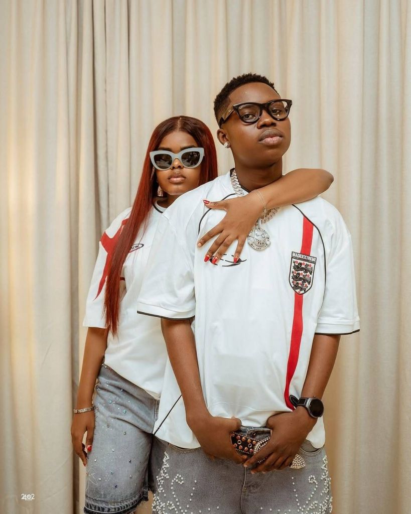 Lovebirds Peller and Jarvis pose affectionately in matching white jerseys with red accents and the number 16, accessorized with sunglasses, jewelry, and stylish denim shorts adorned with pearl embellishments, set against a backdrop of beige curtains.