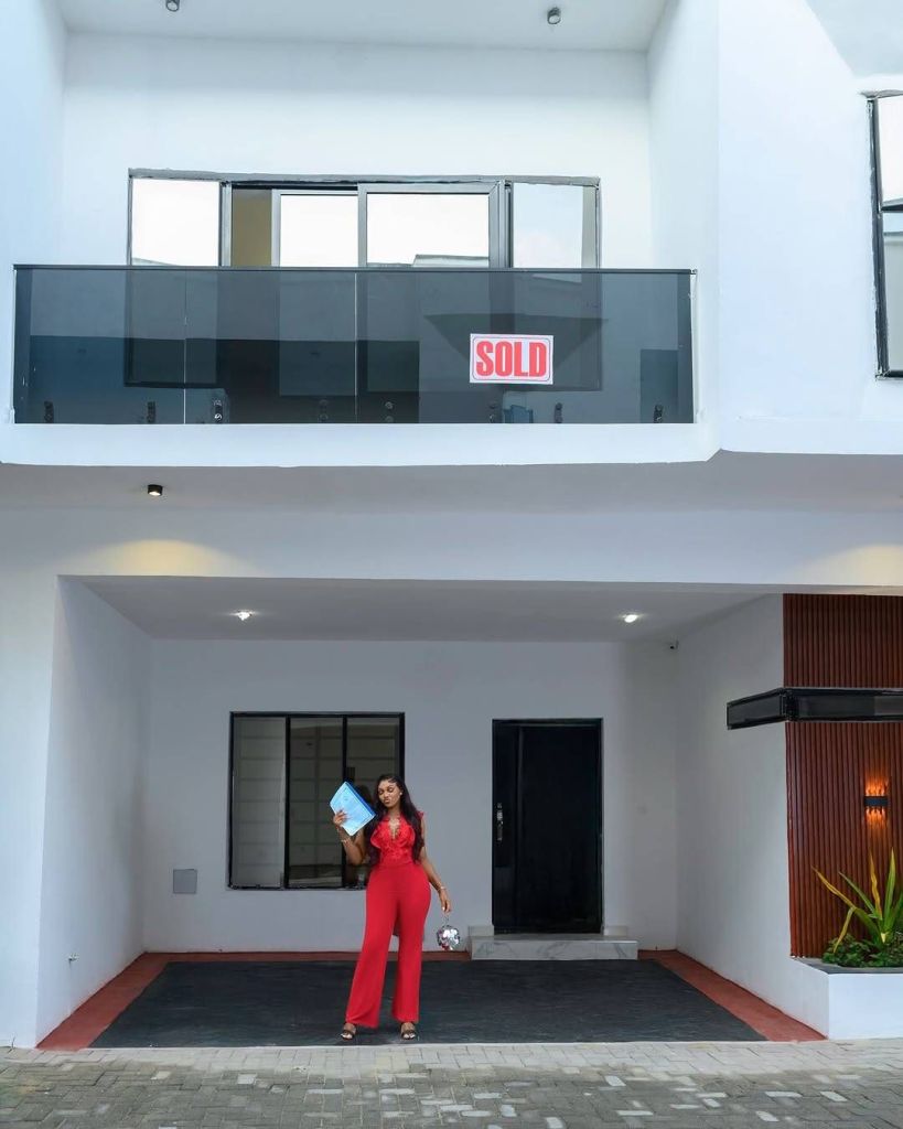 BBNaija star Chomzy stands proudly in front of her newly gifted house, dressed in a striking red jumpsuit. She holds a blue folder in one hand and a small bag in the other, exuding confidence. The modern two-story house features a clean white facade, a glass balcony with a bold "SOLD" sign, and a spacious garage area with a paved driveway, accented by lush green plants.