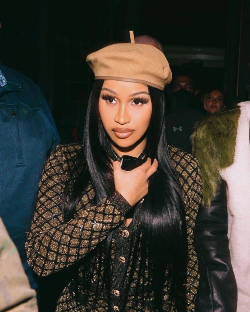 Rapper Cardi B is captured in a stylish moment, wearing a beige beret and a shimmering black jacket with a diamond-patterned design. Her long, sleek black hair cascades over her shoulders as she poses with a hand near her neck, accessorized with a choker. The background features a dimly lit indoor setting with other people, including one in a green furry jacket, adding to the lively atmosphere.