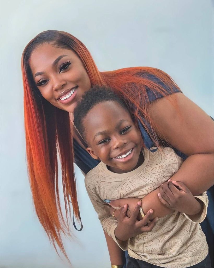 Demure, with long, vibrant red hair, embraces her son against a light background. She wears a dark top, while her son is dressed in a beige long-sleeve shirt with a subtle pattern, both sharing a warm, affectionate moment.