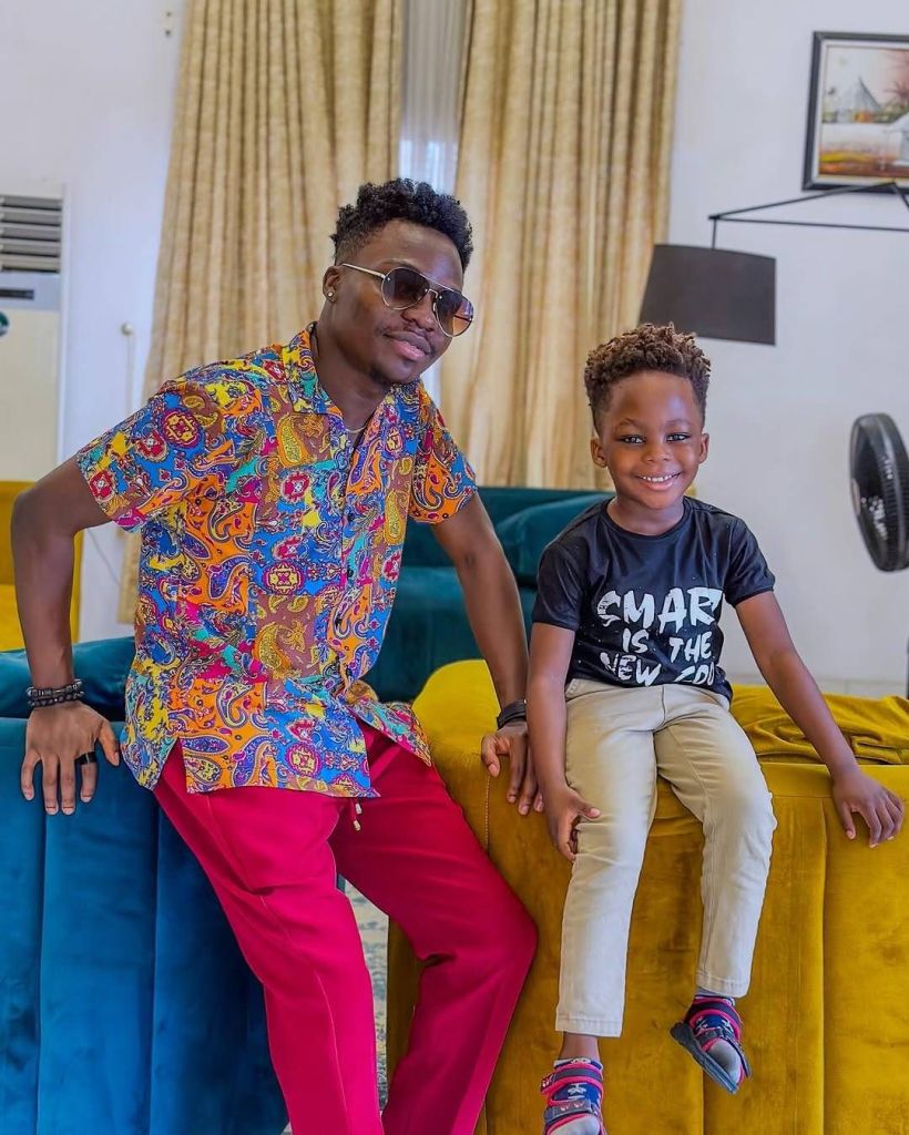 Temiloluwa, dressed in a vibrant multicolored shirt and bright red pants, sits on a blue velvet couch alongside his son. The young boy, wearing a black "SMART IS THE NEW COOL" t-shirt and beige pants, smiles brightly while seated on a yellow cushion. The cozy living room features beige curtains, a modern lamp, and a framed picture on the wall, creating a warm and inviting atmosphere.