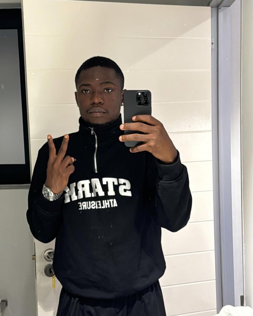 Shank Comics, is taking a selfie in front of a mirror. He has short black hair and is wearing a black hoodie with white text that reads "STAR" and "ATHLEISURE" underneath, although the text appears reversed due to the mirror's reflection. He is also wearing black pants and a silver watch on his left wrist. The man is holding up a peace sign with his left hand and his phone in his right hand. The background features a white wall with a door and a window to the left.