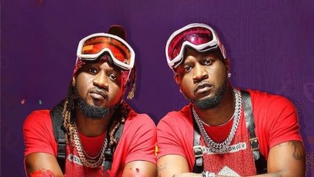 Two individuals, Peter and Paul of the defunct music duo Psquare, are seen wearing matching red outfits, goggles, and heavy gold chains against a vibrant purple background.
