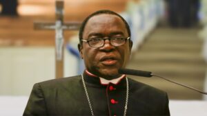 Bishop Kukah, dressed in black clerical attire with red trim and a pectoral cross, speaks at a podium with a microphone in a church setting, with a crucifix visible in the blurred background.