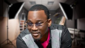 Fuji singer Pasuma, dressed in a black shirt, pink undershirt, and checkered vest, poses in a studio with photography equipment and a blurred background.