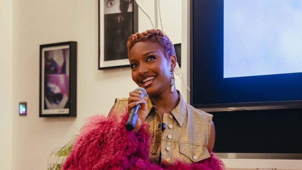 Ayra Starr, smiling and holding a microphone, stands in front of a screen during a presentation. She wears a gold jacket and a fluffy pink shawl, with framed artwork on the wall in the background.