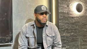BBNaija star Frodd sits against a sleek stone wall with a circular light fixture, wearing a black cap with "DTS" emblazoned on it and tinted sunglasses. He sports a light gray denim jacket over a black shirt, exuding a casual yet stylish vibe. The modern setting includes a subtle vertical light strip, adding to the contemporary ambiance.