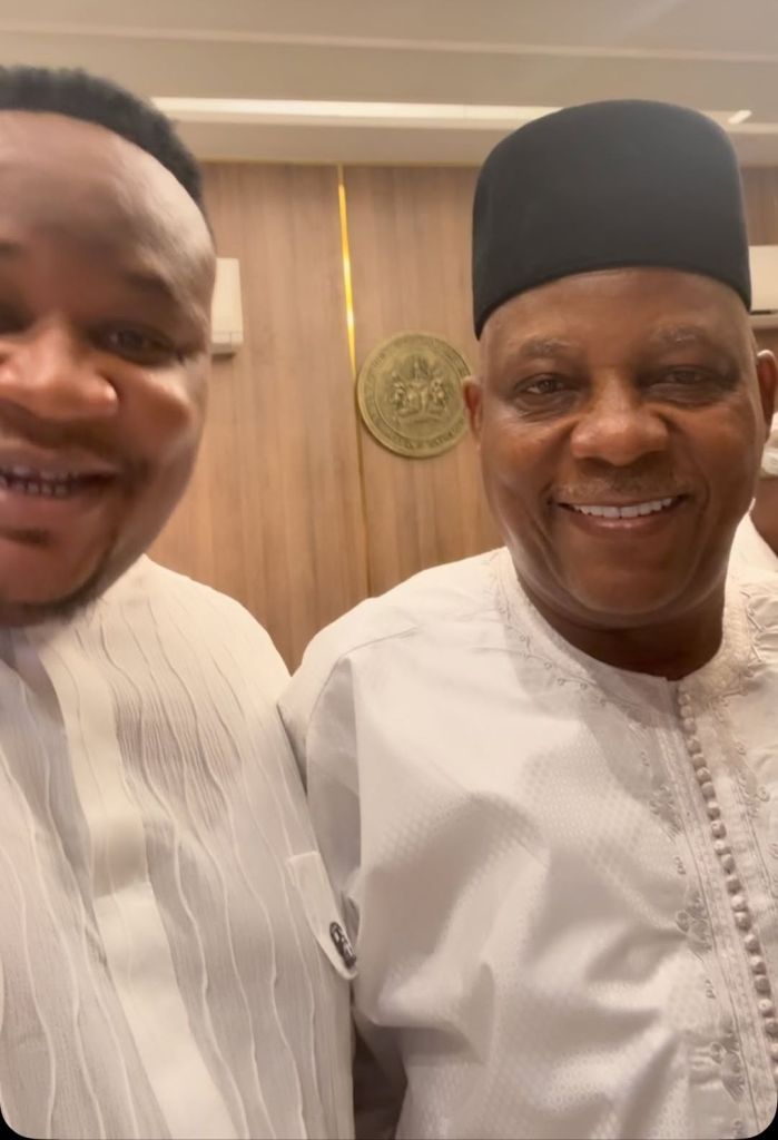 Cubana Chief Priest and Vice President Shettima smile for a selfie in a room with a chandelier, wooden paneling, and a coat of arms on the wall, both dressed in white traditional attire.