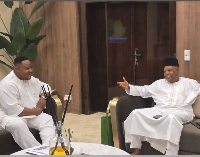 Cubana Chief Priest and Vice President Shettima sit on couches in a luxurious room with wooden paneling and a potted plant, engaged in conversation, with a glass table holding a diffuser and documents between them.