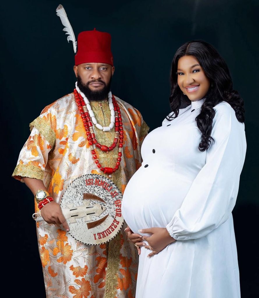 Actor Yul Edochie stands beside his second wife, Judy Austin, who is visibly pregnant. Yul is dressed in a vibrant orange and gold traditional outfit, adorned with red beaded necklaces and a red cap with a feather. He holds a ceremonial fan. Judy wears a flowing white dress, smiling warmly while gently holding her baby bump.