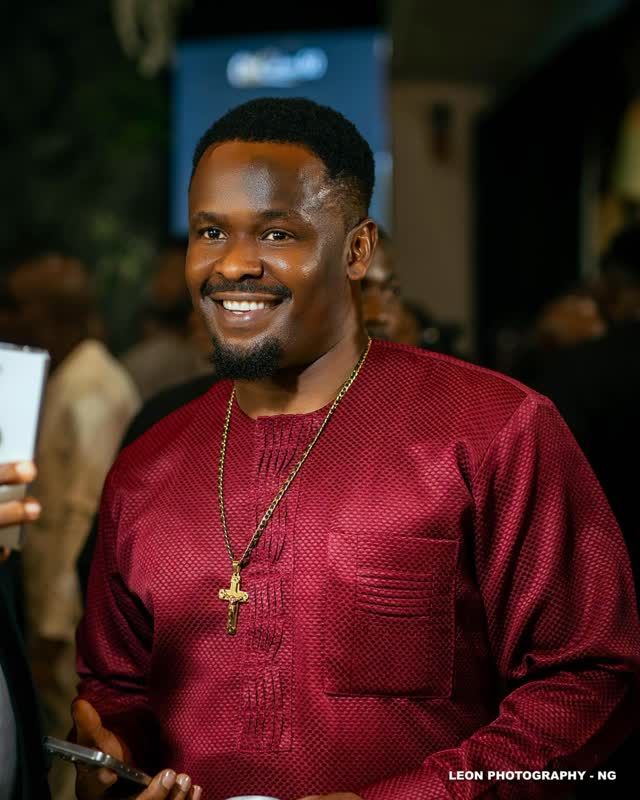 Zubby Michael smiles warmly in a stylish red traditional outfit, accessorized with a gold chain and cross pendant. He holds a phone and a cup, standing among a crowd at an event.