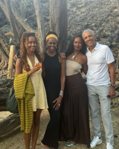 A group of four people, including Barack Obama, Michelle Obama, and their two daughters, stand together outdoors near trees and rocky terrain. They are dressed in casual summer attire, with one daughter holding a drink and another wearing a colorful shawl, all smiling for the photo.