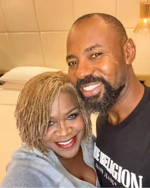 Linc Edochie stands beside Yinka in a softly lit bedroom, wearing a black “True Religion Brand Jeans” t-shirt. Behind them, a bed with white pillows and a beige headboard adds a sense of intimacy to the setting, highlighting a quiet moment between the actor and his estranged wife.