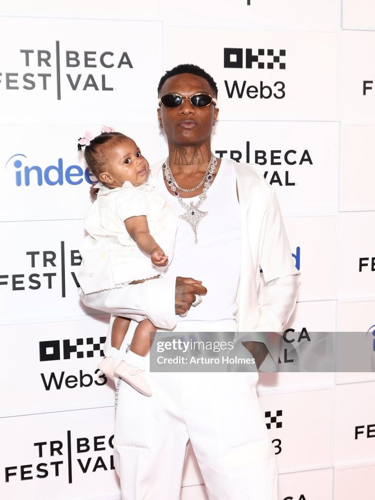 Nigerian singer Wizkid is seen at the premiere of his new documentary, “Wizkid: Long Live Lagos,” at Tribeca Festival, holding his 6-month-old daughter Morayo. Wizkid sports a stylish white outfit with sunglasses and multiple layered necklaces, while Morayo is dressed in a matching white dress adorned with a pink hair accessory and soft pink shoes. The backdrop features "Tribeca Festival," "Web3," and "Indeed" logos.