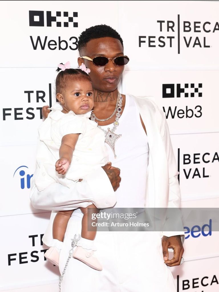 Nigerian singer Wizkid appears at the premiere of his new documentary, “Wizkid: Long Live Lagos,” at Tribeca Festival, holding his 6-month-old daughter Morayo. Wizkid is dressed in a sleek white ensemble with sunglasses and an array of dazzling necklaces, including a prominent star pendant. Morayo wears a matching white dress with a pink hair accessory and soft pink shoes, complementing her father’s style. The backdrop features "Tribeca Festival," "Web3," and "Indeed" logos.
