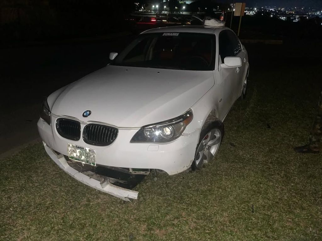 A wrecked white BMW with a damaged front end and Lagos license plate rests on grass at night, with city lights visible in the background and "DUNAMIS" written on the windshield.