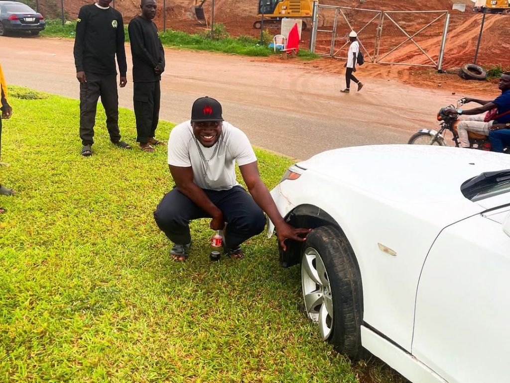BBNaija Ifiemi Angel Awotarigha is seen crouching beside a white car with a damaged tire, smiling in a white shirt and black cap, with others standing nearby on a grassy area near a construction site.
