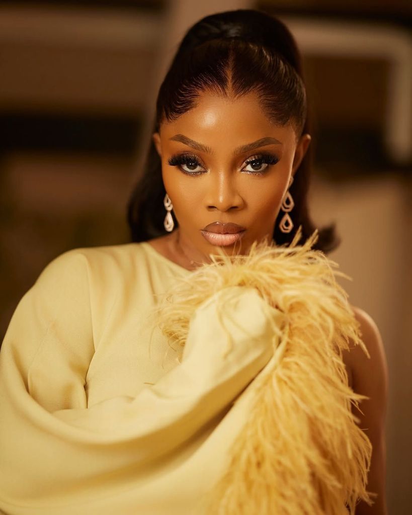 Toke Makinwa wearing a stylish yellow dress with feathered accents, accessorized with elegant dangling earrings, poses confidently with a sleek updo against a warm, neutral background.