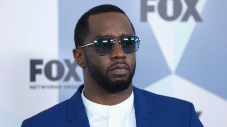 Diddy, dressed in a blue suit and sunglasses, stands in front of a backdrop featuring the FOX logo.