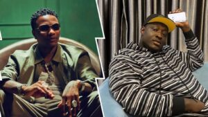 Singer Wizkid appears seated in a green jacket layered over a white shirt, accessorized with bold jewelry, while skitmaker Caterefe lounges on a blue couch in a striped hoodie and cap, focused on his phone. Contrasting backdrops—green and grey—frame each figure, highlighting their individual styles.
