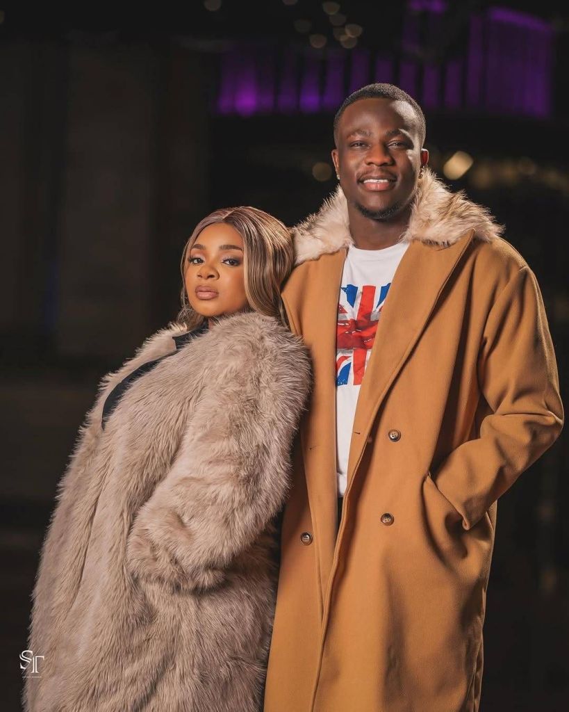 BBNaija star Queen Mercy and her husband David are featured in a stylish moment, dressed in luxurious fur coats. Queen Mercy sports a light-colored fur coat over a black outfit, while David wears a tan coat with a fur-lined collar over a white shirt adorned with a Union Jack design. They pose confidently against a backdrop with purple lighting and a blurred indoor setting.