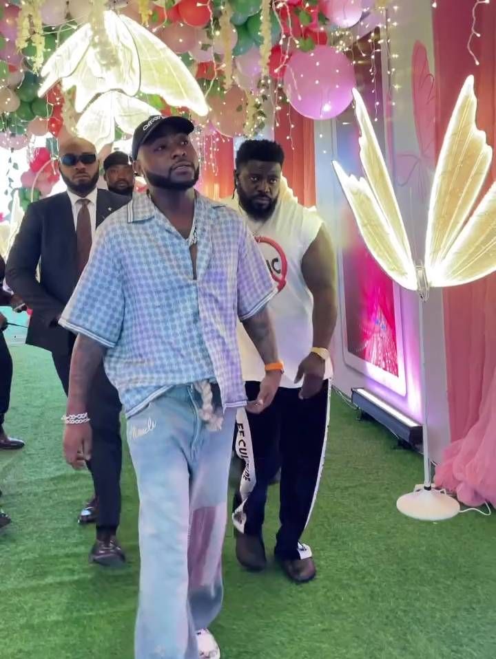 A vibrant scene features singer Davido walking confidently alongside his security personnel on a green carpet. The backdrop is adorned with pink balloons, floral decorations, and illuminated butterfly wings, creating a festive atmosphere. Davido sports a checkered shirt and custom jeans, while his security team follows in suits and casual attire, ensuring a lively yet secure celebration.