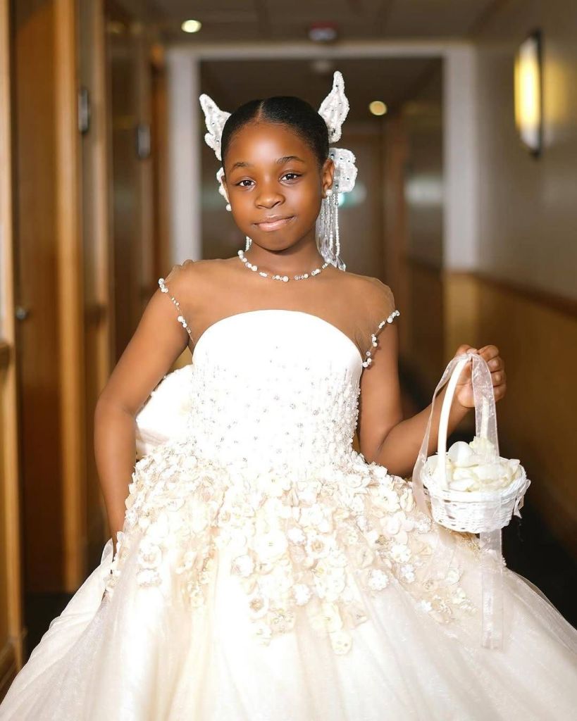 A charming moment captures Davido's first child, Imade, dressed in an elegant white gown adorned with floral embellishments and pearl accents. She stands in a softly lit hallway, holding a small white basket filled with flowers, her hair decorated with delicate butterfly accessories, radiating a fairy-tale charm for a special occasion.