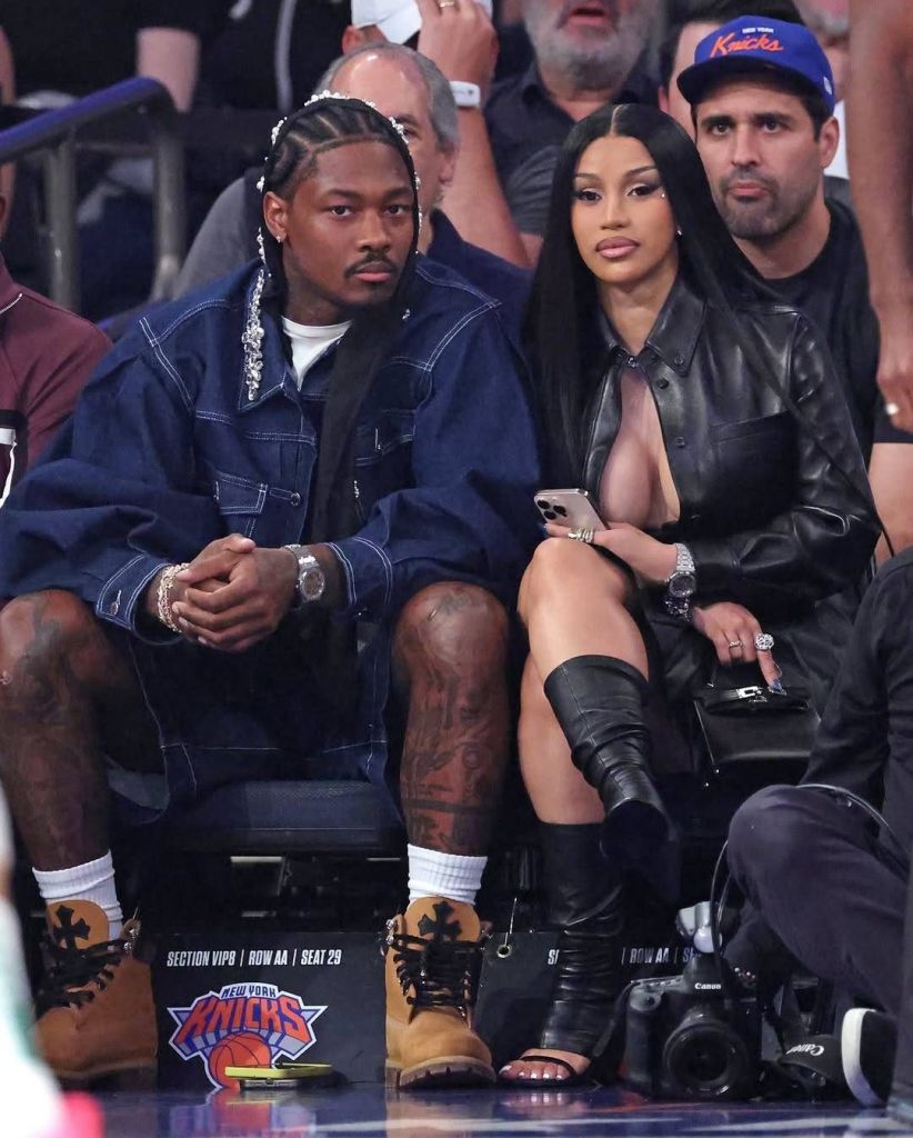 Cardi B and Stefon Diggs sit courtside at a New York Knicks game in the VIP section, Row AA, Seats 29 and 30. Cardi B wears a black leather outfit with thigh-high boots, holding a smartphone and a small black bag, while Stefon Diggs sports a denim jacket, shorts, and tan boots, his tattooed legs visible. A Canon camera rests on the floor nearby, and the crowd in the background watches the game.