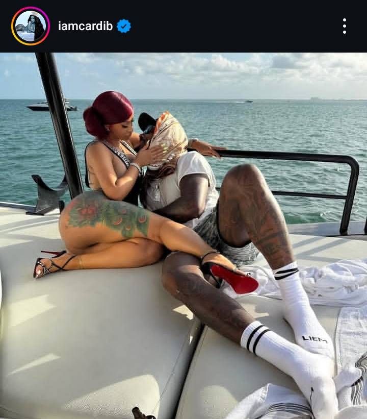 Cardi B and Stefon Diggs relax on a yacht, lounging on a cushioned deck with the ocean in the background. Cardi B, with red hair tied in a bun, wears a black bikini and red high heels, her peacock tattoo visible on her hip, as she leans in for a kiss. Stefon Diggs, dressed in a white shirt and shorts, has his tattooed legs stretched out, wearing white socks with "LIEM" written on them. Another boat is seen in the distance under a partly cloudy sky. The Instagram handle "iamcardib" is displayed at the top.