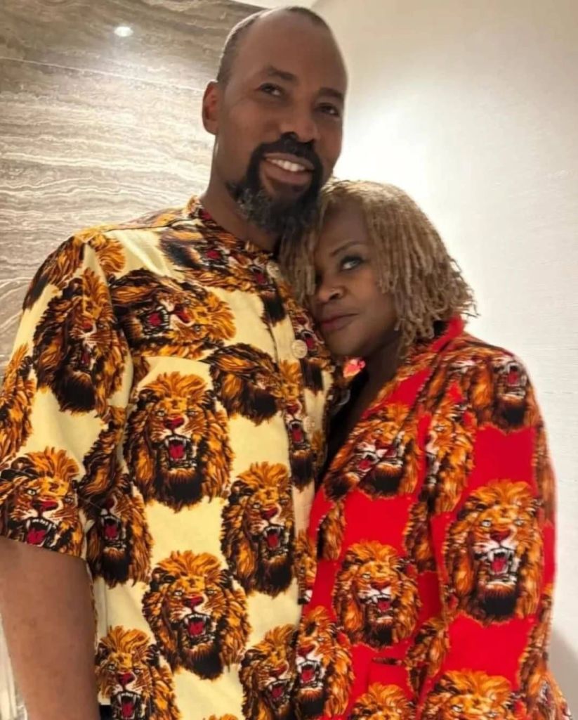 Actor Linc Edochie stands close to his new wife, both dressed in striking outfits featuring bold lion patterns. He wears a cream-colored shirt covered in roaring lion faces, while she sports a vibrant red robe with the same fierce design. The background showcases a modern interior with a marble wall and soft lighting, highlighting their coordinated and eye-catching style.