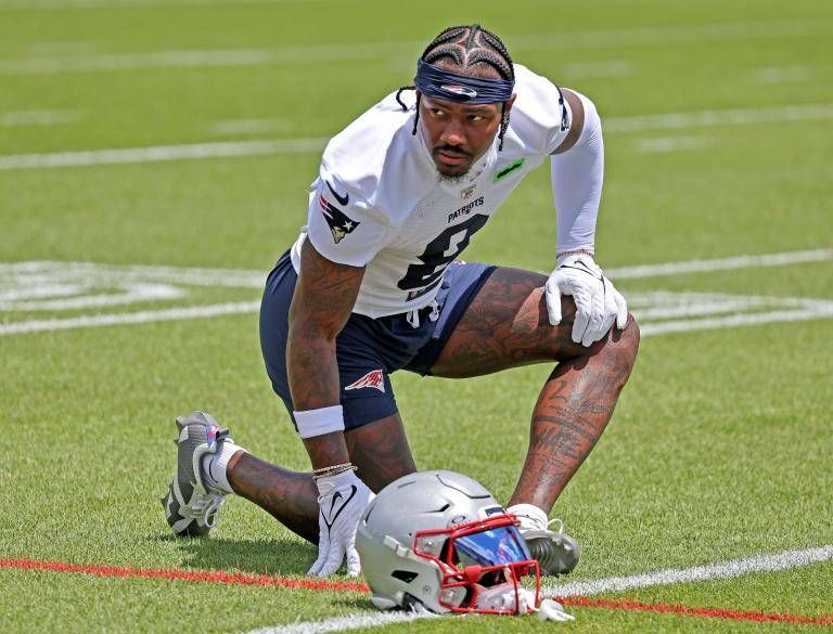 Cardi B's new boyfriend, Stefon Diggs, a football player for the Patriots, captured on the field in a white jersey with the number 8, kneeling beside his helmet. He wears a dark headband, blue shorts, and white gloves, showcasing his tattooed legs on the green turf near a red sideline.