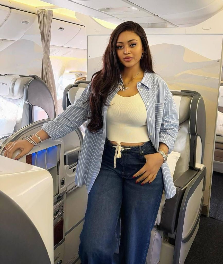 Actress Regina Daniels stands in an airplane cabin, featuring long, straight hair and wearing a light blue striped shirt over a cream top, paired with blue jeans. She accessorizes with a pendant necklace, bracelets, and a watch, with one hand resting on a seat's storage compartment. The modern interior includes plush seats and a window with a scenic view in the background.