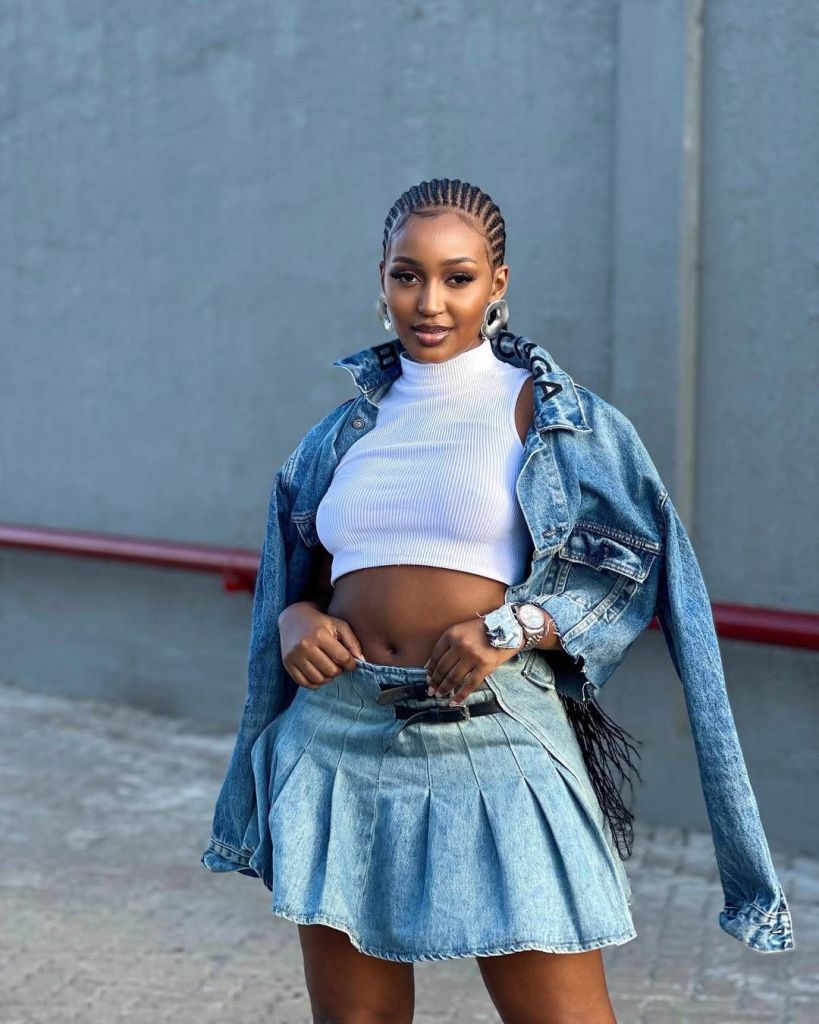 Karen Bujulu, ex-girlfriend of Tanzanian singer Juma Jux, poses confidently against a concrete wall with a red pipe in the background. She rocks a stylish denim-on-denim look, pairing a light-washed pleated mini skirt with a matching oversized jacket. Underneath, a white ribbed crop top accentuates her midriff, while her neatly braided hairstyle is complemented by hoop earrings and a wristwatch, adding a touch of modern flair to her casual ensemble.