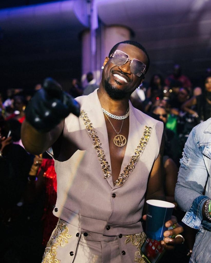 Mr. P stands out in a lively crowd, wearing a stylish beige sleeveless outfit adorned with intricate gold embroidery and embellishments along the edges. He accessorizes with a bold chain necklace featuring a circular pendant, large sunglasses, and a black glove on one hand. Holding a cup, he exudes confidence with a pointed gesture, set against a vibrant backdrop with purple lighting and a bustling audience.