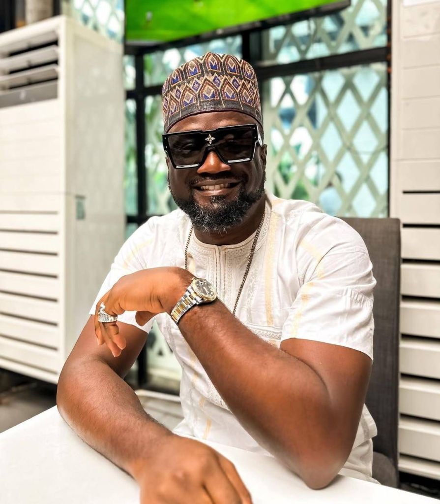 Jude, Mr. P's brother, is seated in a well-lit space, dressed in a traditional white outfit with subtle embroidery, paired with a patterned cap. He accessorizes with a wristwatch, a ring, and a chain necklace, resting his arms on a white table. The background features a geometric-patterned window and a green element, adding a modern touch to the setting.