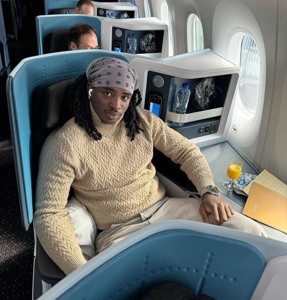 Nigerian skitmaker Lord Lamba relaxes in a luxurious airplane seat, dressed in a beige cable-knit sweater, light trousers, and a gray bandana with white patterns. His long braided hair flows over his shoulders, and he wears wireless earbuds and a sleek watch. A glass of orange juice, a menu, and a bottle of water rest on the tray table beside him, with plush blue seats and airplane windows visible in the background, suggesting a first-class travel experience.